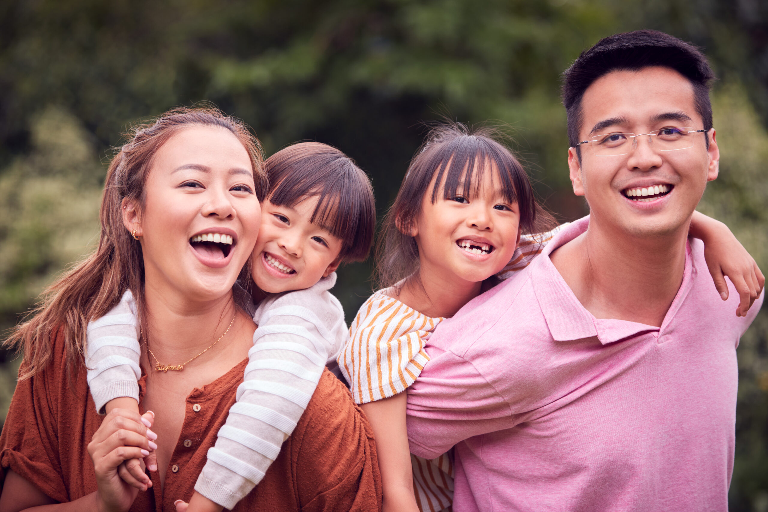 Portrait Of Smiling Asian Family Outdoors With Parents Giving Children Piggybacks